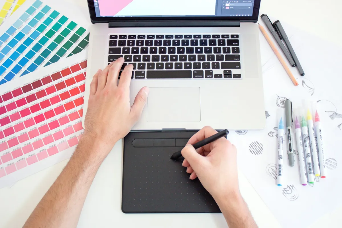 Digital artist using a graphics pen tablet and laptop with design software, colour swatches and sketches