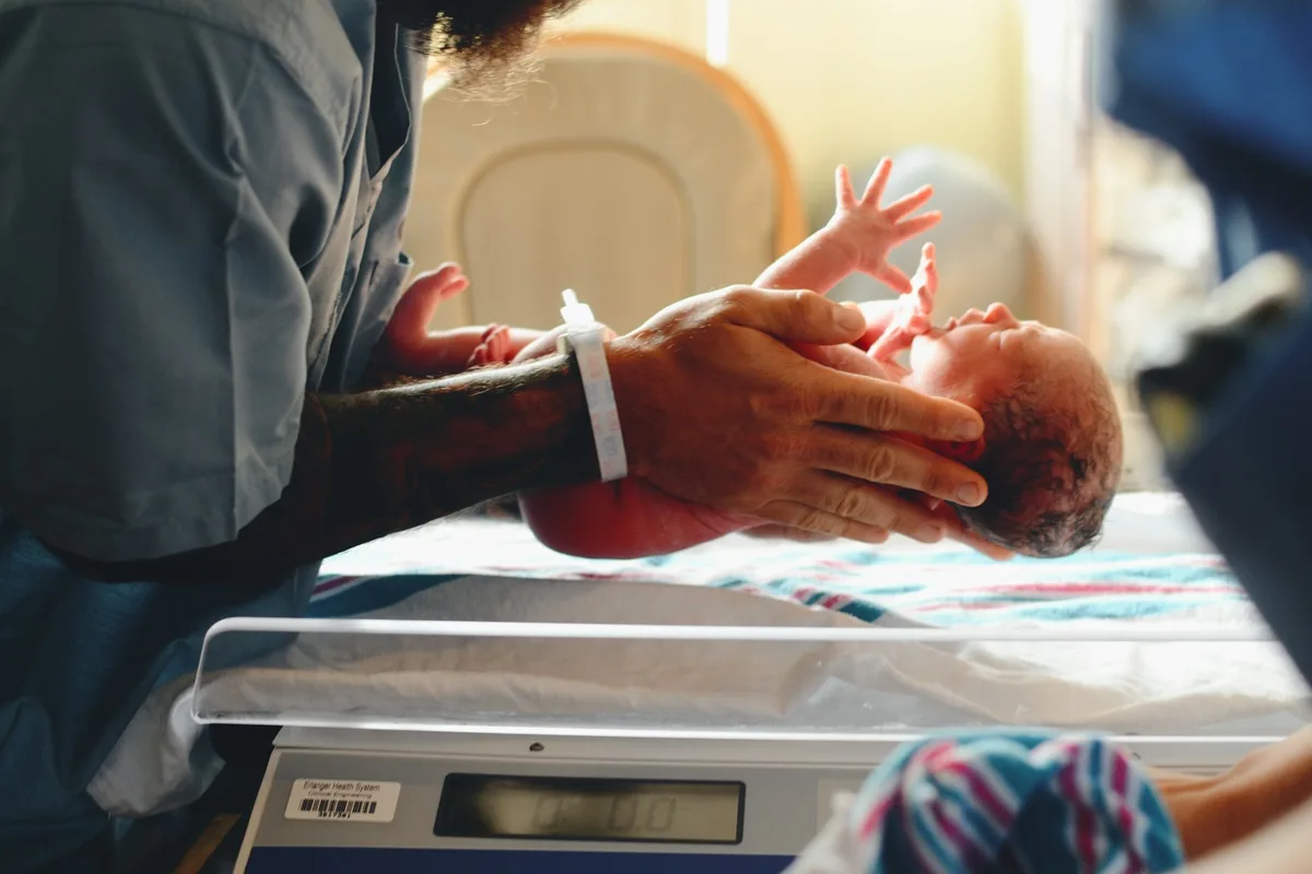 UK midwife smiling beside a newborn baby being held by its mother in a hospital ward