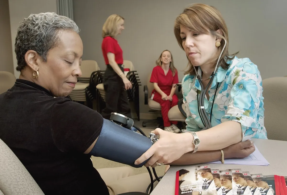 UK nurse with a stethoscope measuring a patient's blood pressure in a clinical setting, another nurse in scrubs visible in the background